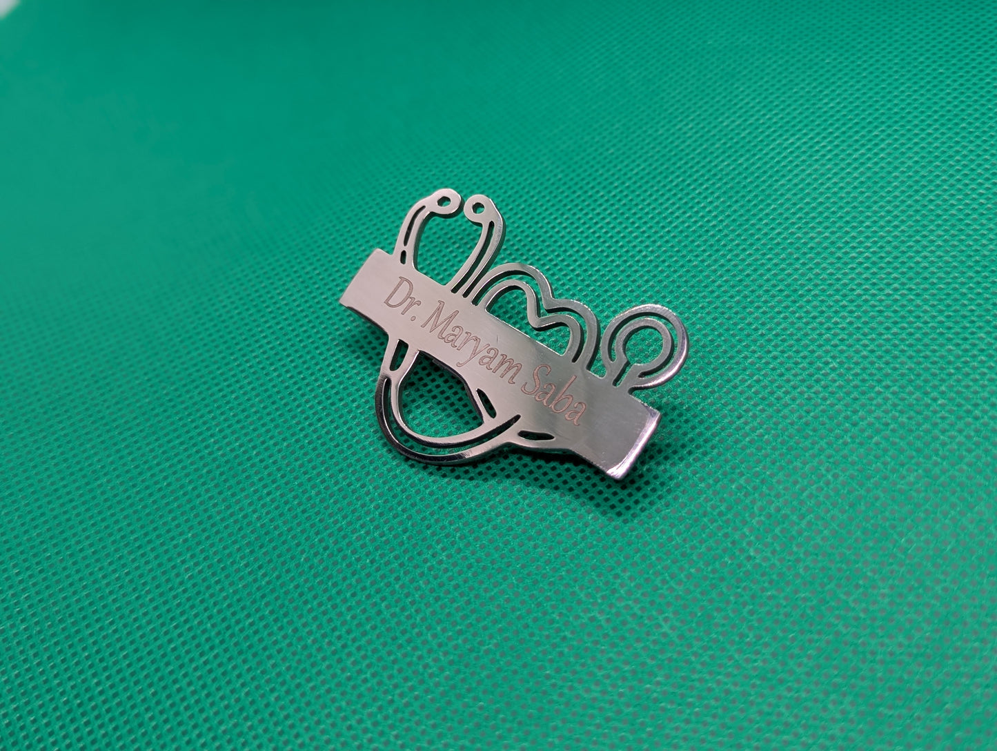 Metallic stethoscope-shaped name badge with engraved text on a green textured background